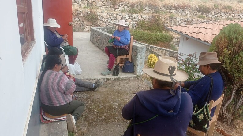 Pu&ograve; includere: Cinque persone sono sedute all'aperto, indossando cappelli e lavorando a maglia. Sono sedute su un patio con un muro di pietra e un tetto di tegole sullo sfondo.
