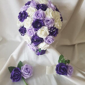 May include: A bridal bouquet and accessories set, featuring artificial roses in shades of purple and cream. The bouquet is a cascade of blooms, with a matching buttonhole and wrist corsage. Rhinestones and pearl accents add a touch of elegance.