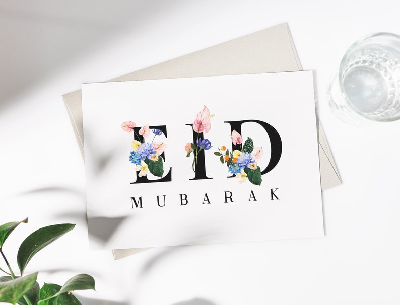 May include: A white card with the words "Eid Mubarak" written in black letters. The letters are decorated with colorful flowers.
