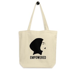May include: A beige tote bag with a black silhouette of a woman wearing a headscarf. The text "EMPOWERED" is printed in black below the silhouette.