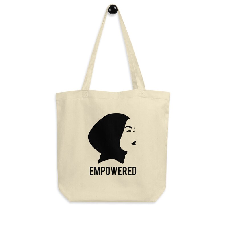 May include: A beige tote bag with a black silhouette of a woman wearing a headscarf. The text "EMPOWERED" is printed in black below the silhouette.
