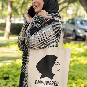 May include: A woman wearing a black and white plaid coat and a black headscarf is holding a beige tote bag with a black silhouette of a woman wearing a headscarf and the word "EMPOWERED" printed on it.