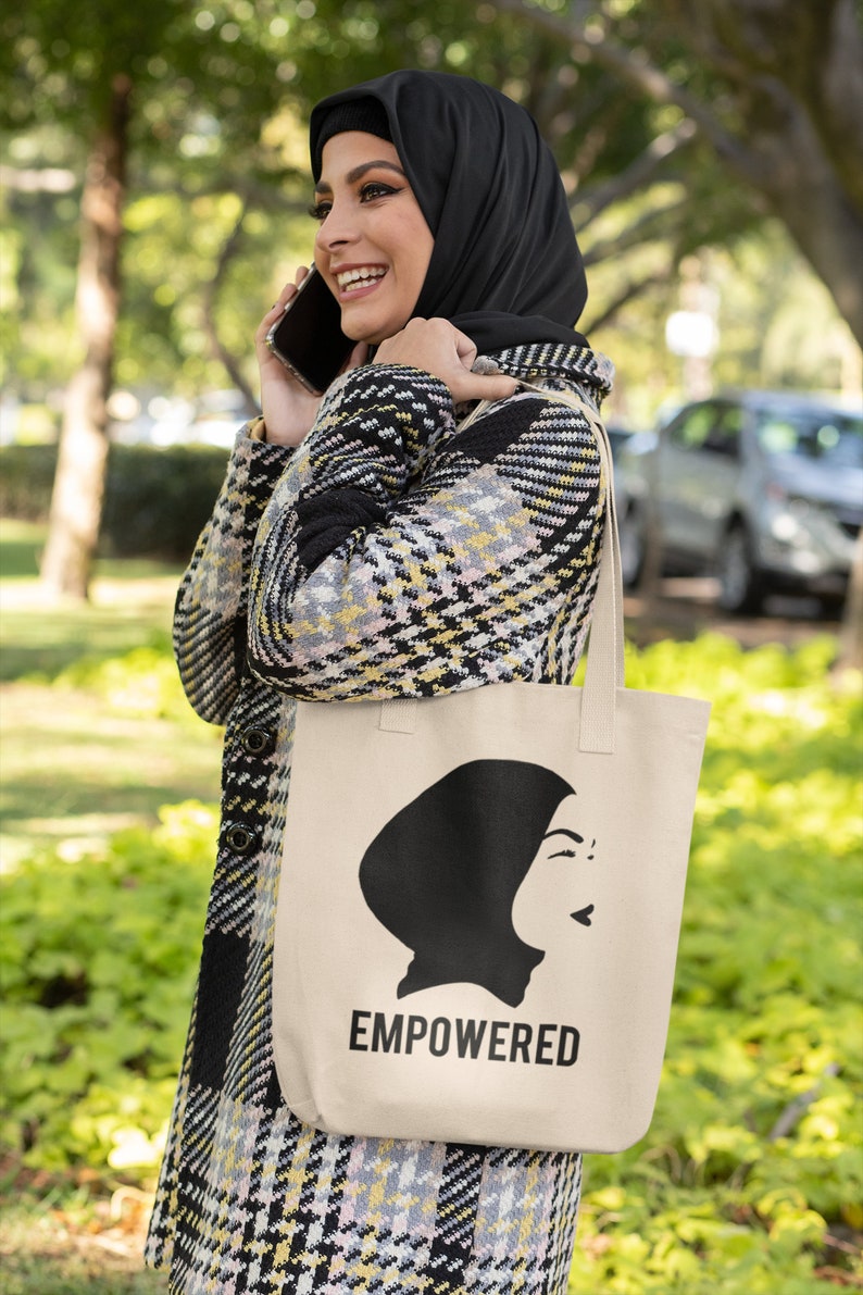 May include: A woman wearing a black and white plaid coat and a black headscarf is holding a beige tote bag with a black silhouette of a woman wearing a headscarf and the word "EMPOWERED" printed on it.