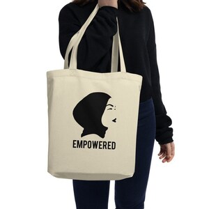 May include: A beige tote bag with a black silhouette of a woman wearing a headscarf. The text "EMPOWERED" is printed in black below the silhouette.