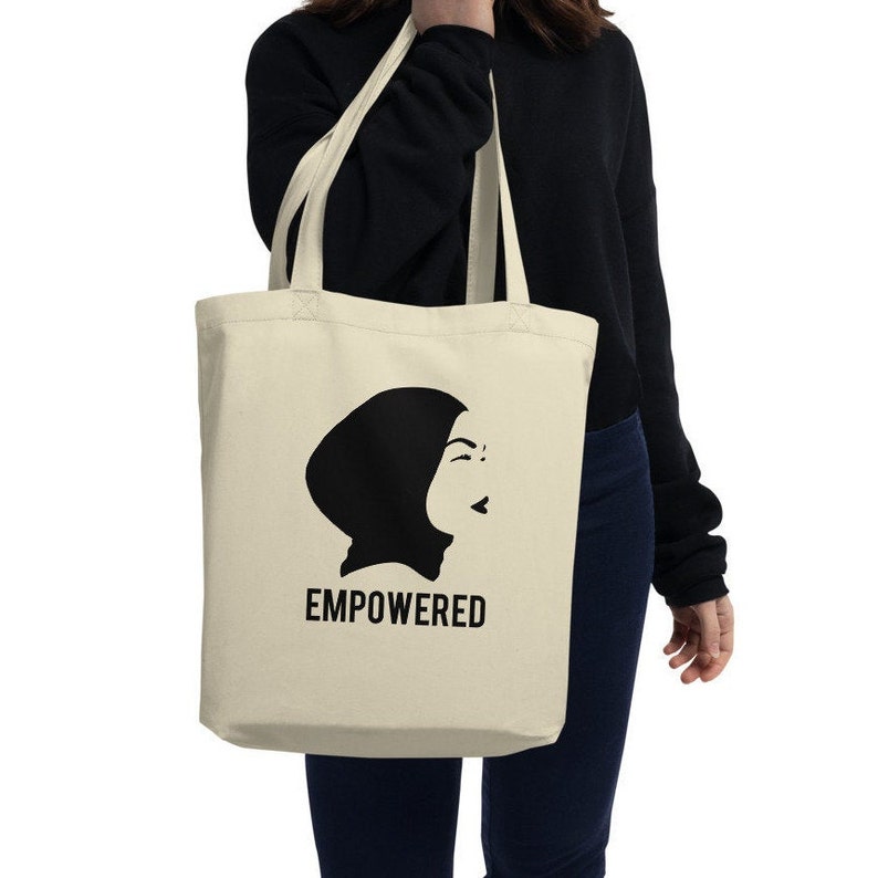 May include: A beige tote bag with a black silhouette of a woman wearing a headscarf. The text "EMPOWERED" is printed in black below the silhouette.