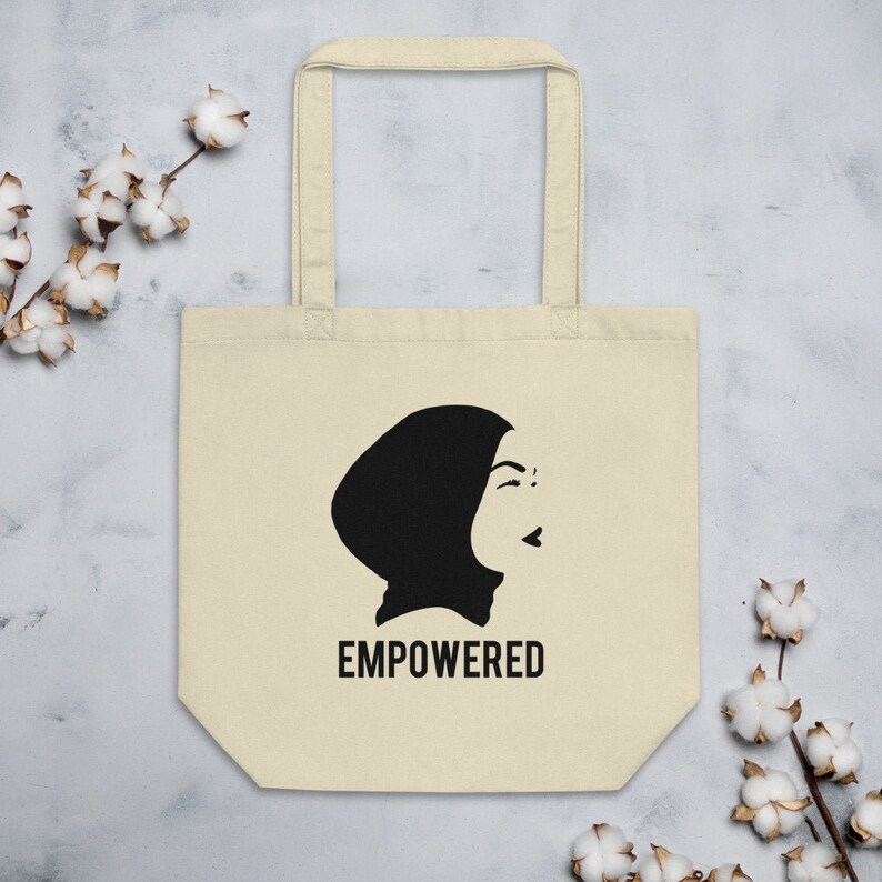 May include: A beige tote bag with black handles and a black silhouette of a woman wearing a headscarf. The text "EMPOWERED" is printed in black below the silhouette.
