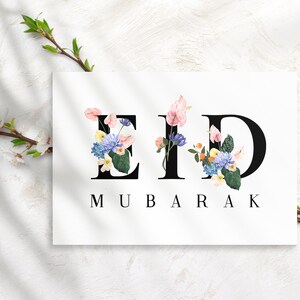 May include: A white card with the words "Eid Mubarak" in black lettering. The letters are decorated with colorful flowers.