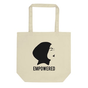 May include: A beige tote bag with a black silhouette of a woman wearing a headscarf. The text "EMPOWERED" is printed in black below the image.
