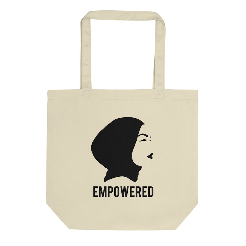 May include: A beige tote bag with a black silhouette of a woman wearing a headscarf. The text "EMPOWERED" is printed in black below the image.