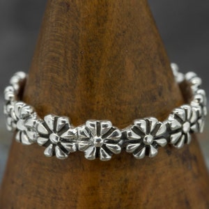 Puede incluir: Un anillo de plata con un delicado diseño floral. El anillo está formado por una serie de pequeñas flores en relieve dispuestas en círculo alrededor de la banda.