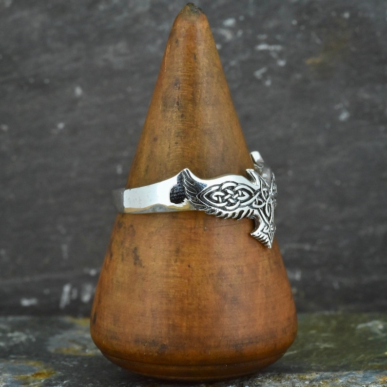 May include: Silver ring with a Celtic knot design in the shape of a bird. The ring is on a wooden display stand.