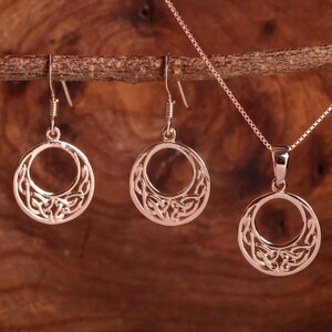 Op de afbeelding: Een ros&eacute;gouden sieradenset met een hangerketting en bijpassende oorbellen. De oorbellen zijn van het druppeltype en de hanger en oorbellen hebben een cirkelvormig ontwerp met een open Keltisch knoopmotief. De sieraden hangen aan een houten tak.