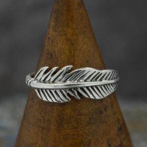 May include: A silver ring featuring a detailed feather design. The ring is displayed on a wooden ring holder.