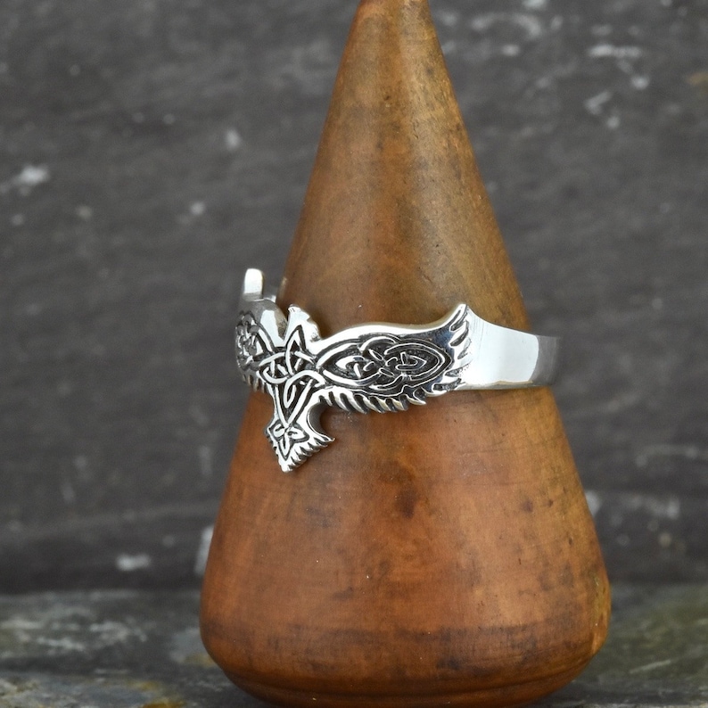 May include: Silver ring with a detailed design of a bird with Celtic knotwork. The ring is displayed on a wooden ring holder.