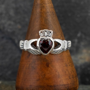 Peut inclure: Une bague Claddagh en argent avec une pierre précieuse rouge en forme de cœur et une couronne sur le dessus. La bague est sur un présentoir en bois.