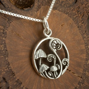 Puede incluir: Un collar con colgante de plata con un dije circular con un diseño de hongos y helechos. El colgante cuelga de una delicada cadena de plata. El dije tiene un diseño detallado inspirado en la naturaleza.