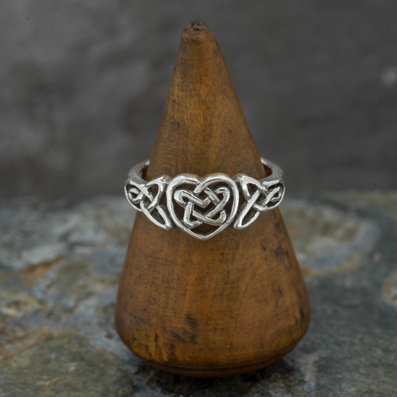 May include: A silver ring with a Celtic heart knot design. The ring features a heart-shaped knot in the center, flanked by intricate knotwork on either side. The ring is displayed on a wooden cone-shaped stand against a mottled gray background.