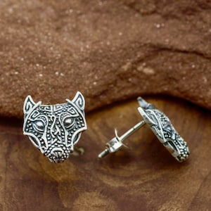 Puede incluir: Un par de pendientes de plata con un diseño detallado de cabeza de lobo. La cara del lobo está tallada intrincadamente con patrones geométricos.
