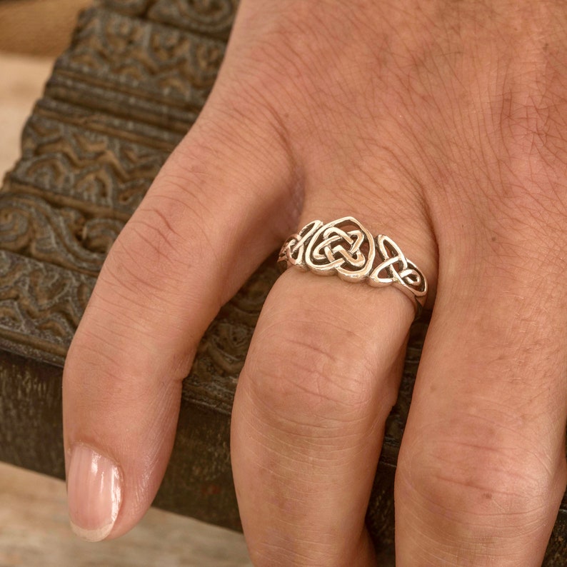 May include: A silver-colored ring featuring a Celtic knot design. The ring is worn on a finger, showcasing the intricate interwoven patterns. The ring's design includes a central heart-shaped knot and other Celtic knot elements.