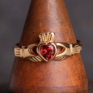 May include: A gold Celtic knot ring with a red heart-shaped gemstone. The ring features a traditional Claddagh design with two hands clasped around a heart.