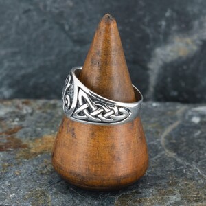 May include: A silver ring with a Celtic knot design. The ring is displayed on a wooden ring holder.