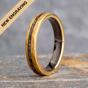 May include: A gold and black wedding band with a hammered finish. The band has a thin black line running around the edge.