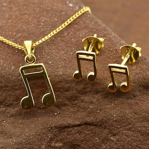 May include: A gold necklace and earrings set featuring a musical note design. The necklace has a chain and a pendant shaped like a musical note. The earrings are also shaped like musical notes and have post backs.