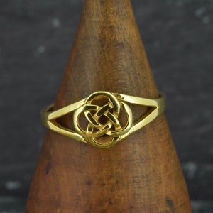 May include: A gold ring with a Celtic knot design. The ring has a central knot and a split band. It is displayed on a wooden stand against a dark background. The ring is a piece of jewellery.