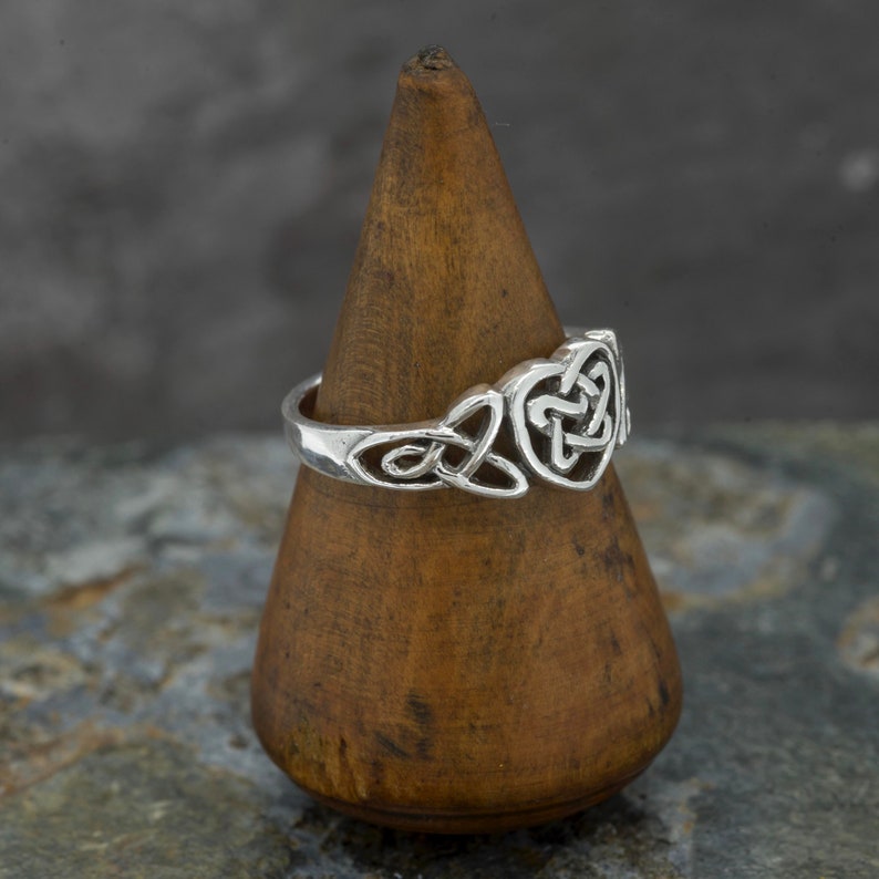 May include: A silver ring with a Celtic knot design. The ring features a heart-shaped knot in the center and other knotwork on the band. The ring is displayed on a wooden cone-shaped stand against a gray background.
