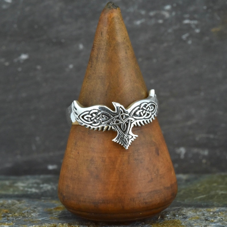 May include: A silver ring with a Celtic knot design featuring a stylized bird. The ring is displayed on a wooden ring holder.