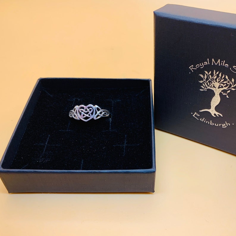 May include: A silver-colored ring with a Celtic heart design, displayed in a black box. The ring is intricately detailed with interwoven patterns. A dark blue box with gold text and a tree logo is visible.