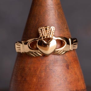 Könnte beinhalten: Ein goldfarbener Claddagh-Ring mit einem Herzen, zwei Händen und einer Krone obenauf.
