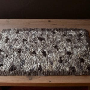 Könnte beinhalten: Ein rechteckiger, handgefertigter Filzteppich mit einem gesprenkelten Muster. Der Teppich besteht aus einer Mischung aus grauen, weißen und dunkelbraunen Wollfasern. Er liegt auf einem Holztisch und zeigt seine Textur.