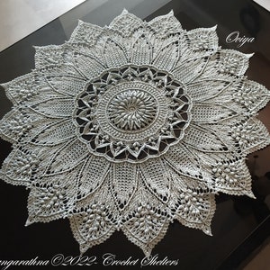 May include: A delicate, light blue crochet doily with a floral design. The doily is laid flat on a dark surface. The text "Ooiya" is visible in the upper right corner of the image. The text "Gangarathna ©2022 Crochet Shelters" is visible in the lower left corner of the image.