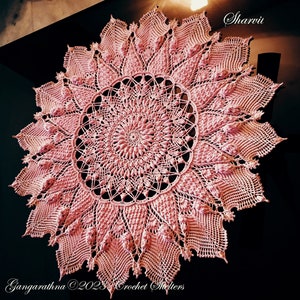 May include: A large, pink crochet doily with a detailed, intricate design. The doily has a circular shape with a central motif and radiating points. The text "Sharvii" and "Gangarathna ©2023 Crochet Shelters" is visible in the image.