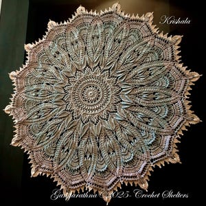Può includere: Un centrino all'uncinetto circolare e dettagliato nei toni del beige, grigio e azzurro. Il design presenta un motivo floreale centrale circondato da intricati motivi a forma di foglia. I testi "Krishala" e "Gangarathna 2025- Crochet Shelters" sono visibili.