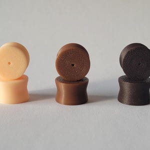 Può includere: Tre paia di dilatatori per orecchie in legno impilati in diverse tonalità di marrone. La parte superiore di ogni dilatatore ha una superficie testurizzata.