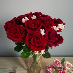 May include: A bouquet of handmade, crocheted red roses with green leaves and small white flowers in a clear glass vase. A few pink and white flowers are on the table next to the vase. The background is a light gray wall.