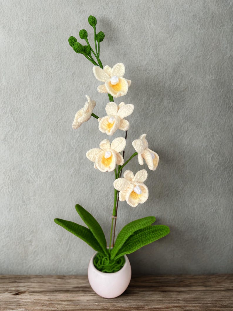 May include: A crocheted orchid plant in a white pot. The off-white flowers have yellow centers and are attached to a green stem. The plant has green leaves and is set against a gray background.