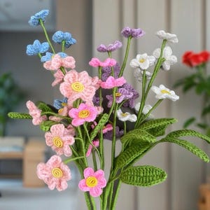 May include: A bouquet of handmade crochet flowers in various colors, including pink, blue, purple, and white. The flowers have yellow centers and green stems and leaves. The bouquet is a craft item.