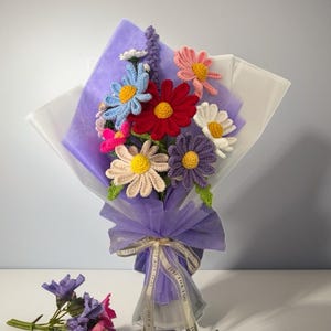 Op de afbeelding: Een boeket van gehaakte bloemen in verschillende kleuren, waaronder roze, rood, paars, blauw en geel, verpakt in paars en wit tissuepapier met een wit lint waarop "Just for you" staat.