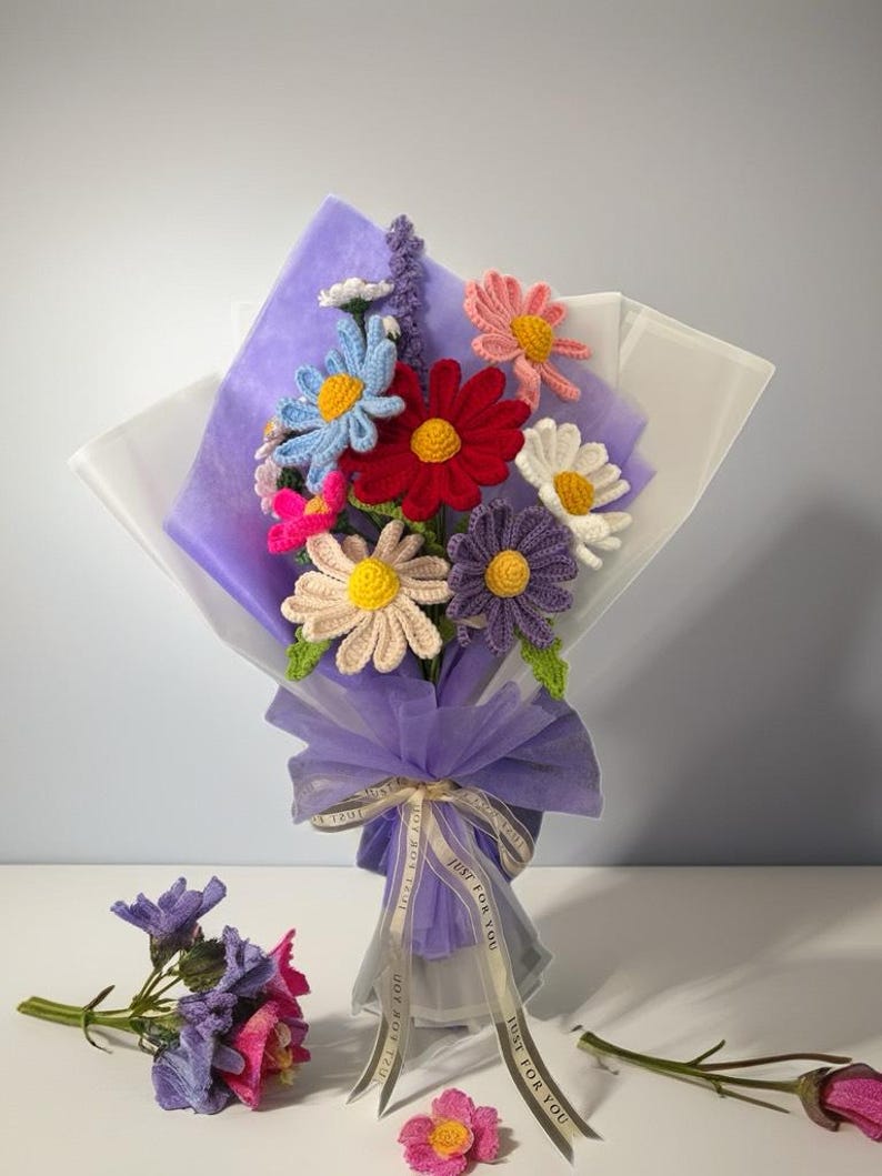 Op de afbeelding: Een boeket van gehaakte bloemen in verschillende kleuren, waaronder roze, rood, paars, blauw en geel, verpakt in paars en wit tissuepapier met een wit lint waarop "Just for you" staat.