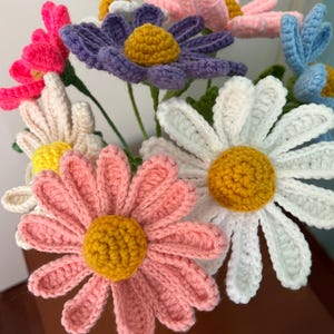 Op de afbeelding: Een boeket van gehaakte bloemen in verschillende kleuren, waaronder roze, wit, paars, blauw en geel. De bloemen zijn in een vaas gerangschikt en hebben een realistisch uiterlijk.