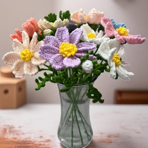 Può includere: Un bouquet di fiori colorati all'uncinetto in un vaso di vetro trasparente. I fiori presentano vari colori, tra cui rosa, bianco, viola e arancione, con centri gialli. Foglie e steli all'uncinetto verdi completano l'arrangiamento floreale fatto a mano.