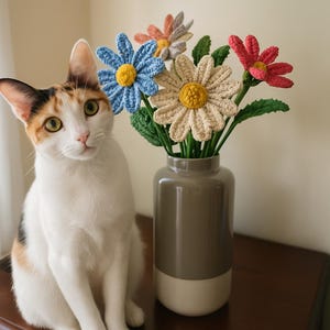 Op de afbeelding: Een wit-oranje kat zit op een houten tafel naast een vaas met gehaakte bloemen. De bloemen zijn blauw, roze en cr&egrave;me met gele centra.