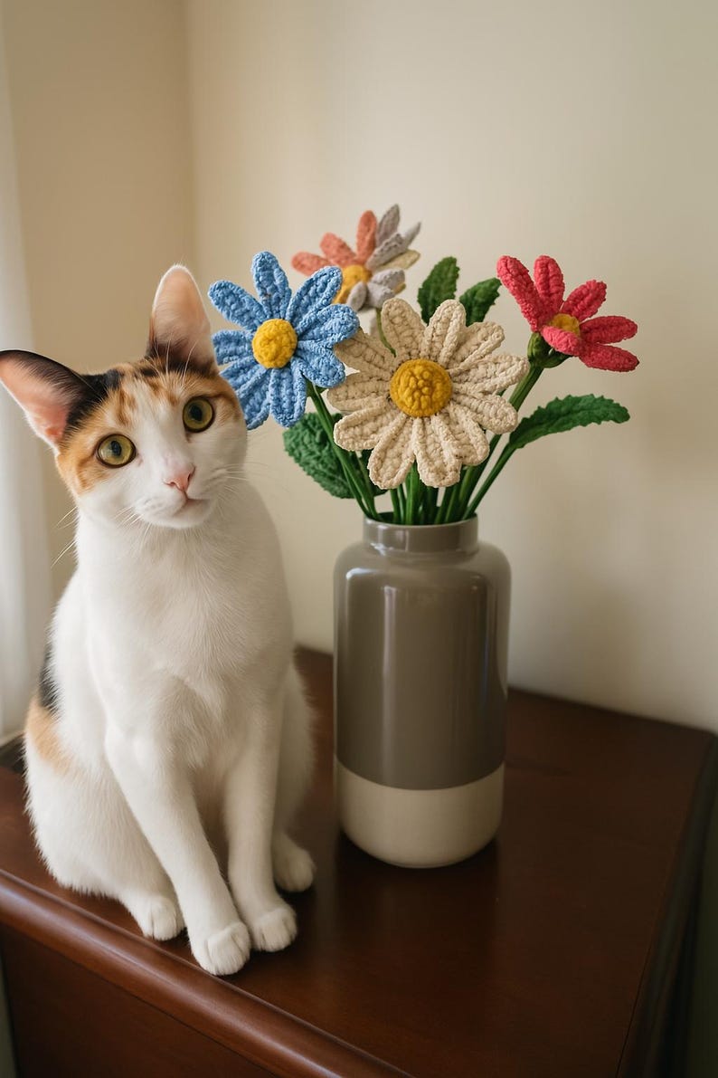 Op de afbeelding: Een wit-oranje kat zit op een houten tafel naast een vaas met gehaakte bloemen. De bloemen zijn blauw, roze en cr&egrave;me met gele centra.