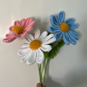 Op de afbeelding: Een boeket van drie gehaakte madeliefjes in roze, wit en blauw met gele centra. De bloemen zijn bevestigd aan groene stelen.