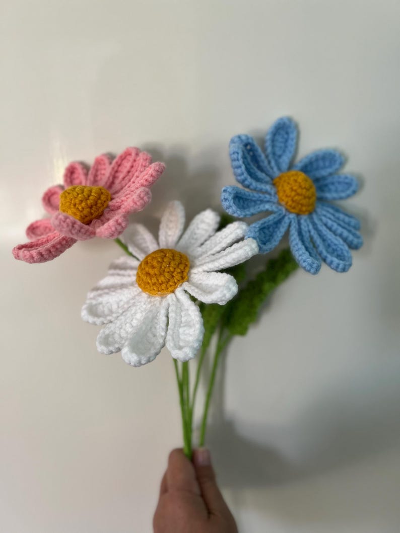 Op de afbeelding: Een boeket van drie gehaakte madeliefjes in roze, wit en blauw met gele centra. De bloemen zijn bevestigd aan groene stelen.