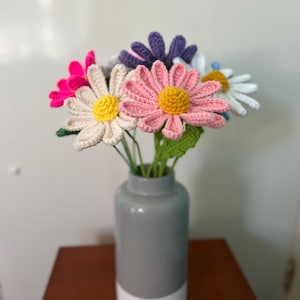Op de afbeelding: Een boeket gehaakte madeliefjes in een grijze en witte vaas. De bloemen zijn roze, wit, paars en geel.