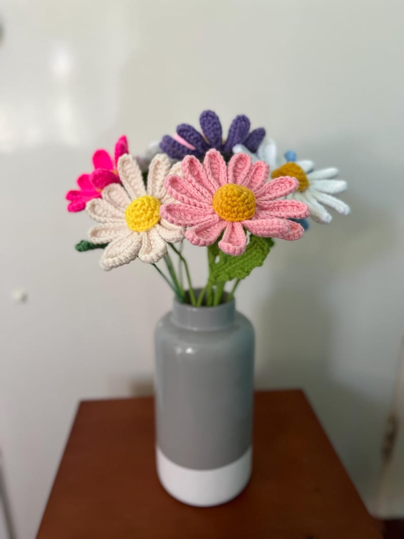 Op de afbeelding: Een boeket gehaakte madeliefjes in een grijze en witte vaas. De bloemen zijn roze, wit, paars en geel.